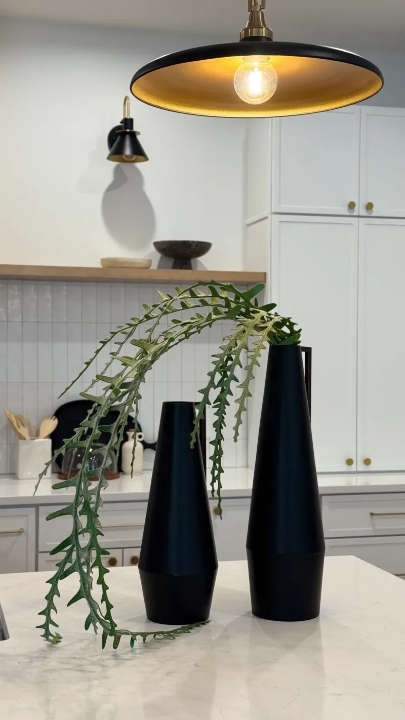 Two black vases with trailing greenery on a marble counter, under a black barn pendant