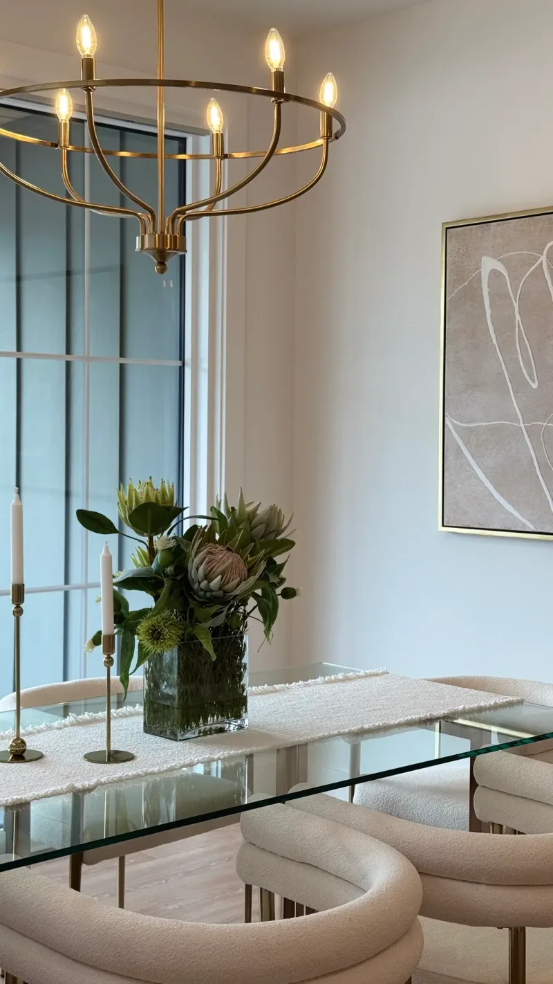 Glass dining table under a brass chandelier, with protea in a clear vase