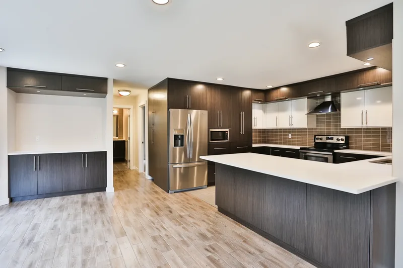 Remodelled kitchen with dark wood-grain and glossy white cabinets, white quartz peninsula, stainless refrigerator and tiled backsplash