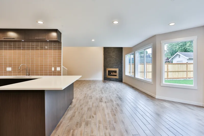 View across the empty great room from the kitchen peninsula toward a stacked-stone fireplace and windows onto the yard