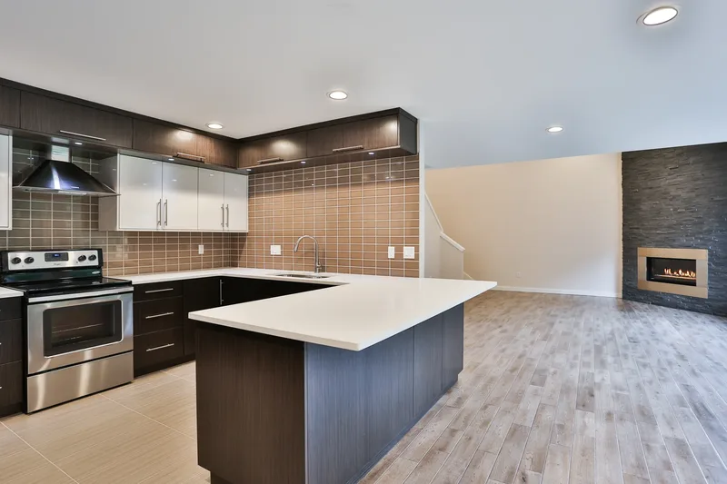 Kitchen from the range side, white gloss uppers above dark base cabinets, quartz peninsula, lit fireplace beyond