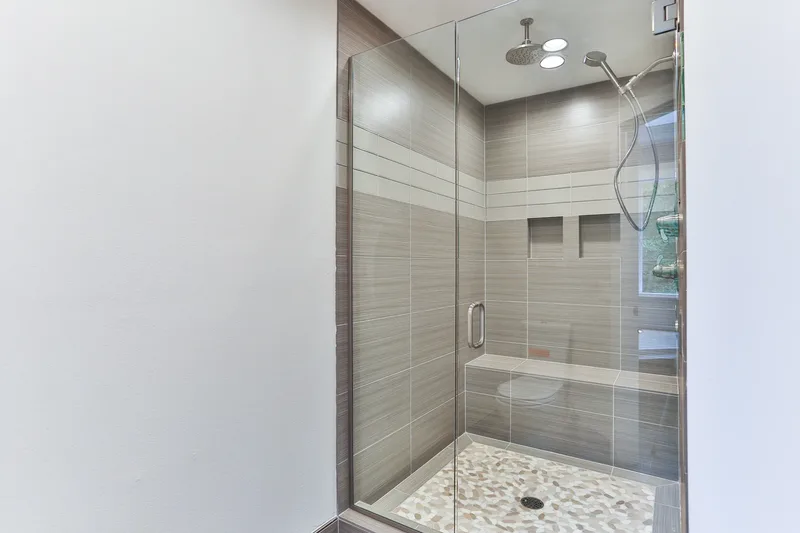 Walk-in shower behind clear glass, striated tile walls, built-in bench, pebble mosaic floor, rain and handheld heads