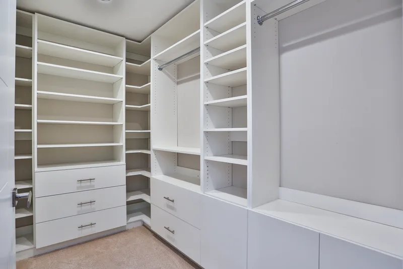 Empty walk-in closet fitted with white shelving, hanging rods, banks of drawers and a low bench