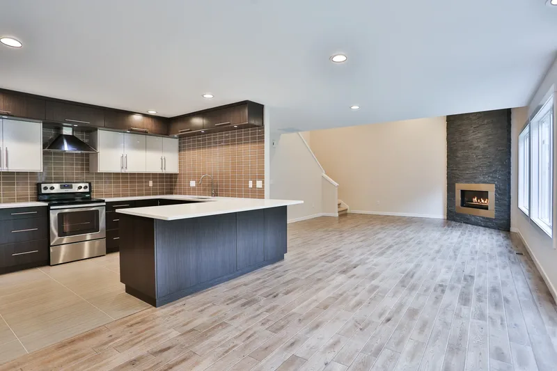 Wide view of the open kitchen and unfurnished living area, peninsula at centre, stone fireplace at right