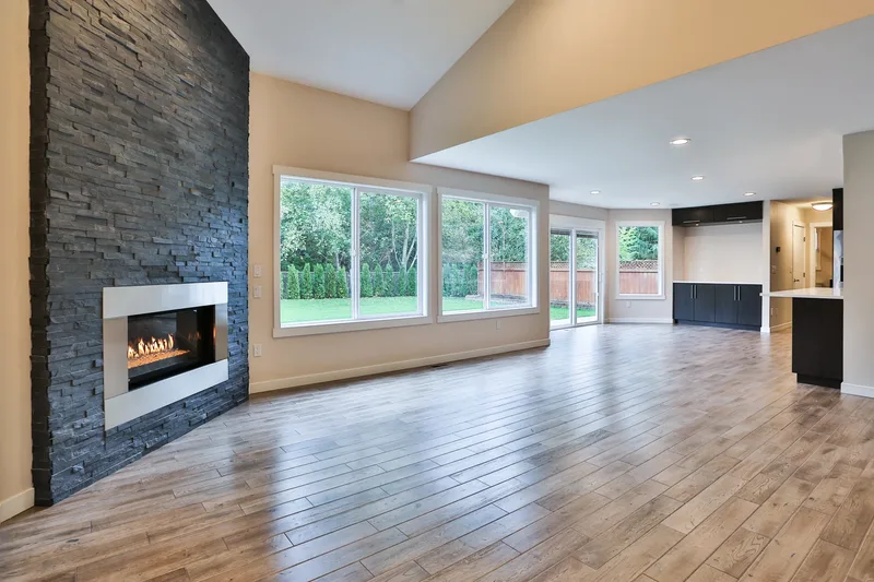 Great room under a vaulted ceiling, lit stone fireplace at left, window wall onto the lawn, kitchen beyond