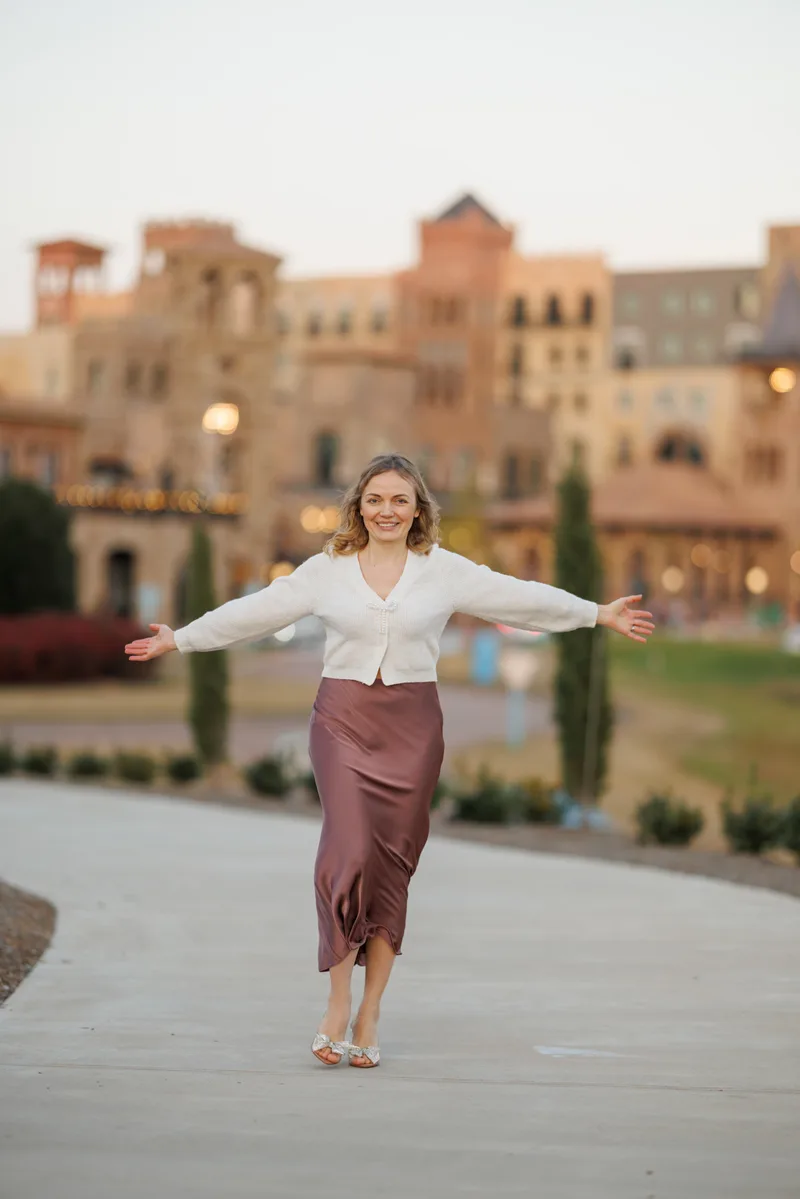 Tatyana walking toward the camera with her arms open, in a cream cardigan and a satin skirt