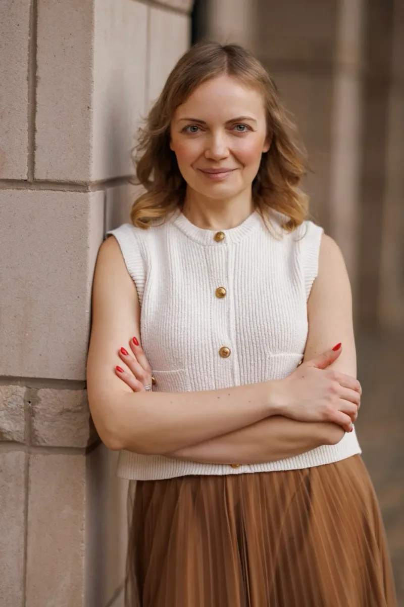 Tatyana in a cream knit vest and tan pleated skirt, against a stone wall