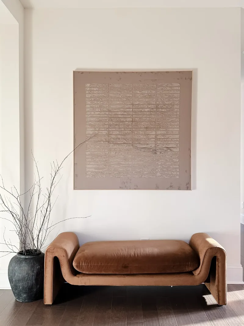 A caramel velvet bench with curved arms below a large square textured canvas, dark branch-filled vase alongside