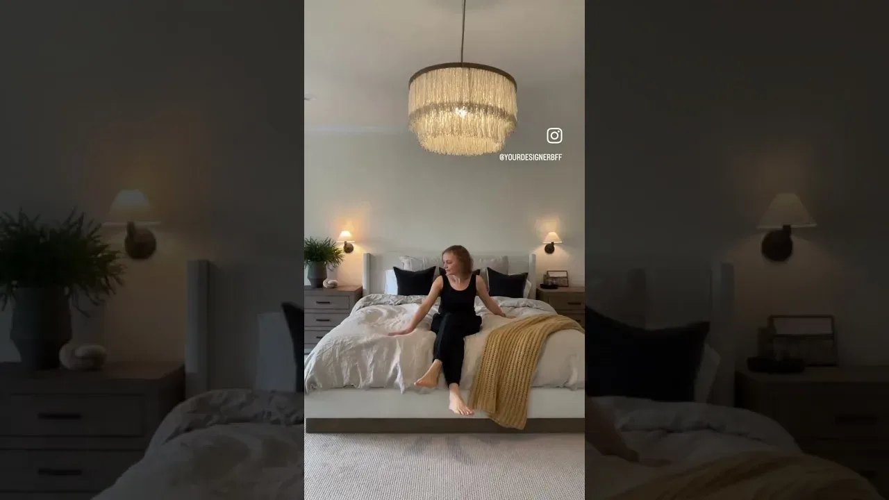 Video still: the layered linen bed with a mustard throw under a fringed drum chandelier