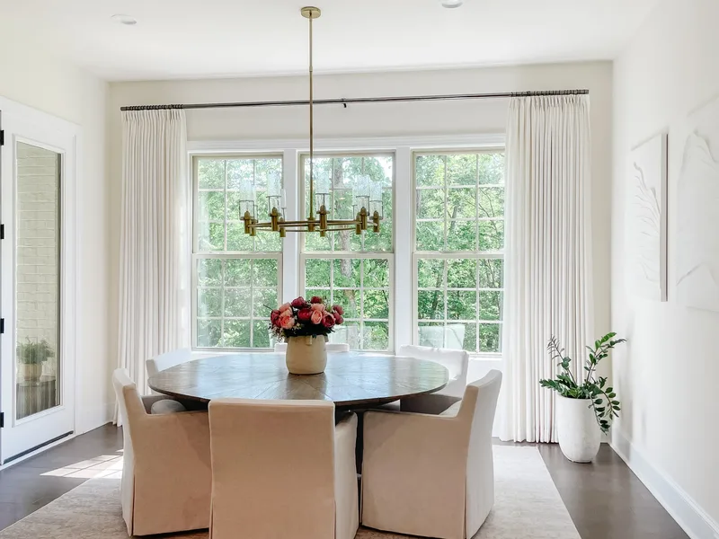 The round table and six slipcovered chairs seen from the doorway, three tall windows and floor-length drapes behind
