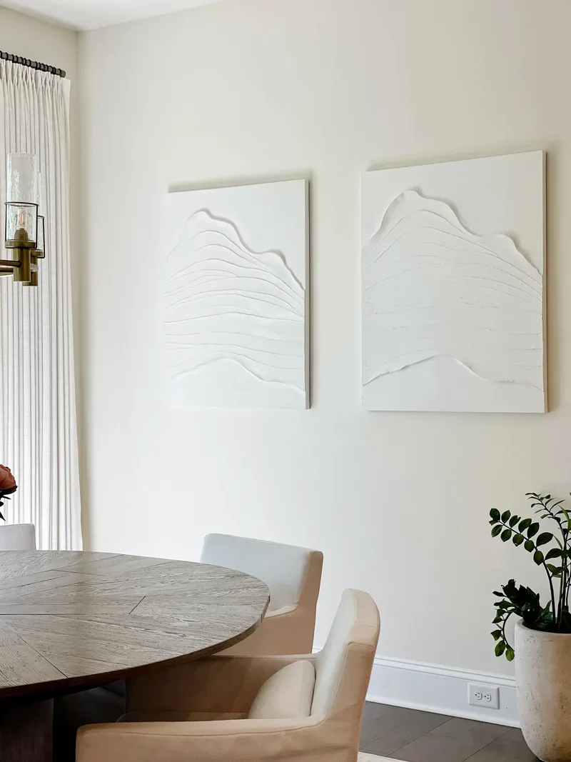 White textured plaster canvases hang on the dining wall above the table edge and a potted plant