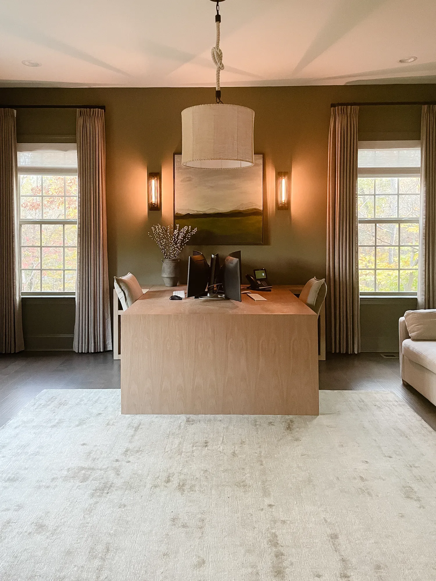 A pale oak waterfall desk with two monitors, set against olive walls between two tall curtained windows